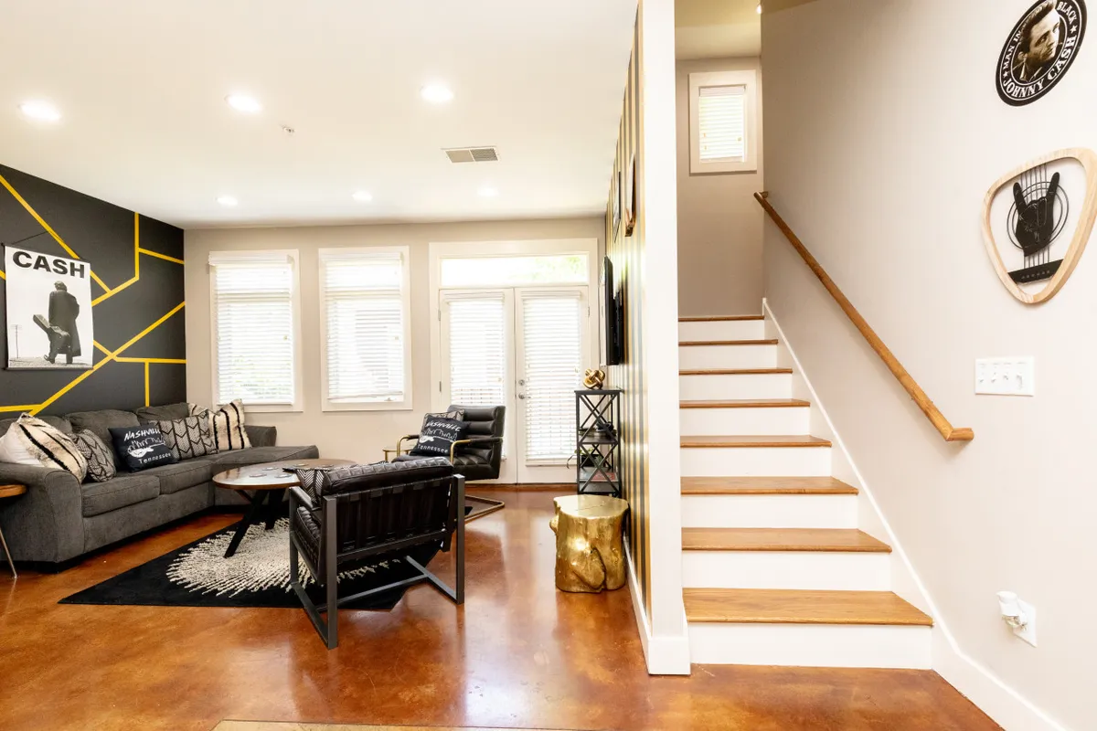 A stylish, comfortable living area showing a sofa with decorative pillows, an elegant gold side table, stairway access, and artistic decor. The design balances comfort and sophistication, offering an ideal space for relaxing or chatting with friends.