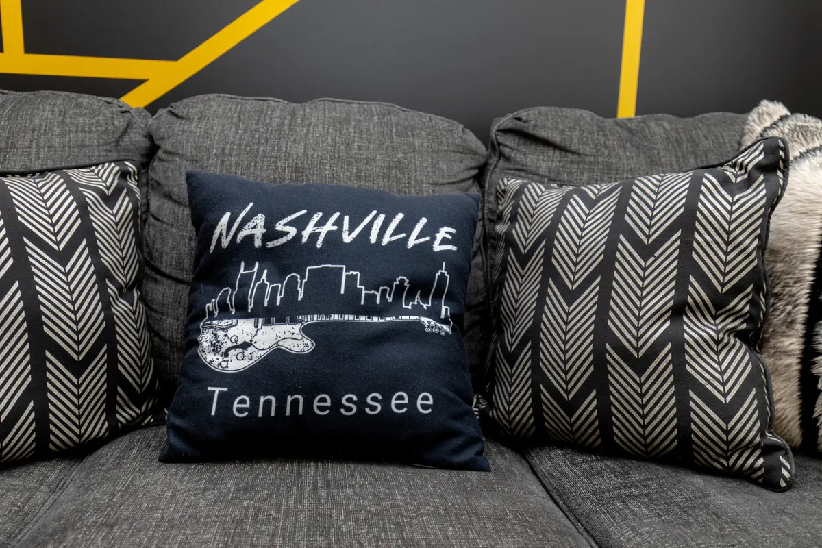 A cozy living area featuring a decorative pillow with a 'Nashville' design, surrounded by textured throws on a comfortable couch. The nostalgic yet inviting mood reinforces the music-themed décor, perfect for relaxing or swapping stories about Music City adventures.