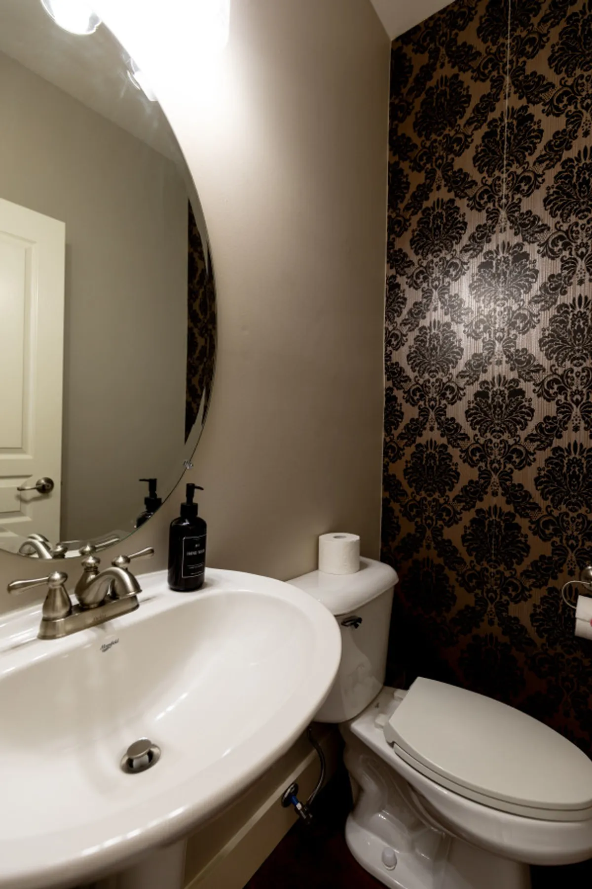 An elegant half bath with a distinctive oval mirror, decorative wallpaper, a cozy sink, and a toilet. The space feels inviting and well-appointed for guests.