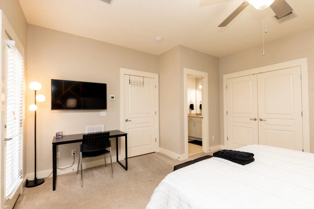 This image shows a serene bedroom with a comfortable bed, a desk with a chair suitable for remote work or study, a television, and a lamp, creating a calm and inviting atmosphere. The space is uncluttered and functional, ideal for both relaxation and productivity.