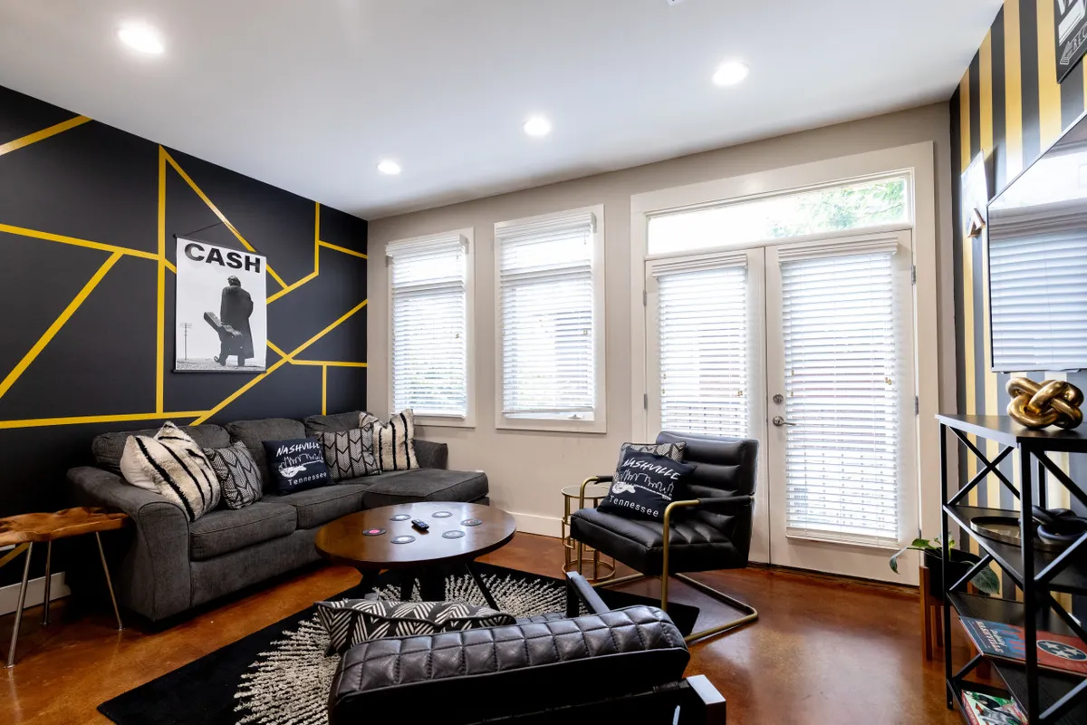 This image features a modern living room with a geometric accent wall, a sofa adorned with decorative pillows, a round coffee table, stylish black chairs, and prominent artwork displaying the word 'CASH'. The décor is bold and directly references the Johnny Cash theme, creating an immediate sense of place.