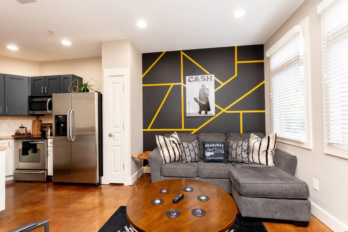 This image captures a cozy, open-plan living area featuring a comfortable sofa, a coffee table, stylish wall art, and a visible kitchen area in the background. The space exudes a welcoming atmosphere, ideal for relaxing after exploring the neighborhood or coming back from a busy day of campus visits.