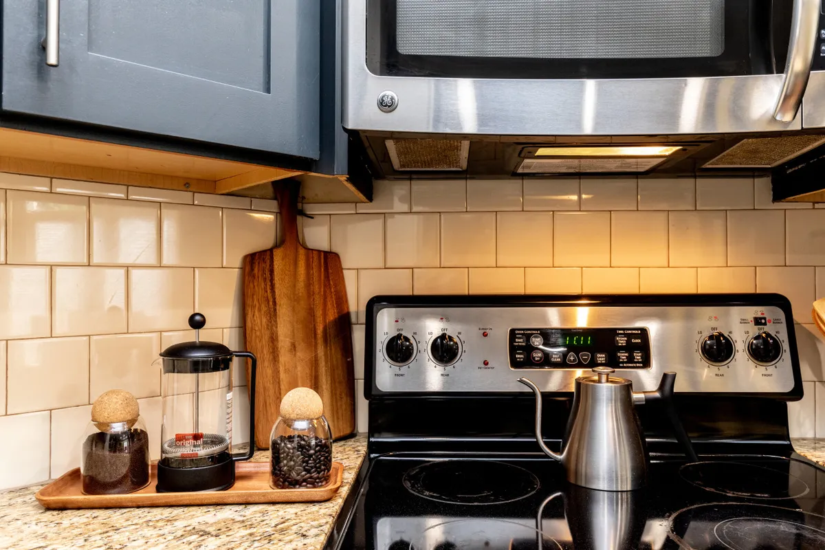 A bright kitchen scene featuring a stainless steel stove, a French press for coffee, a wooden cutting board, and a glass jar filled with coffee beans. The workspace is well-lit and organized, inviting guests to prepare meals and enjoy morning rituals together.