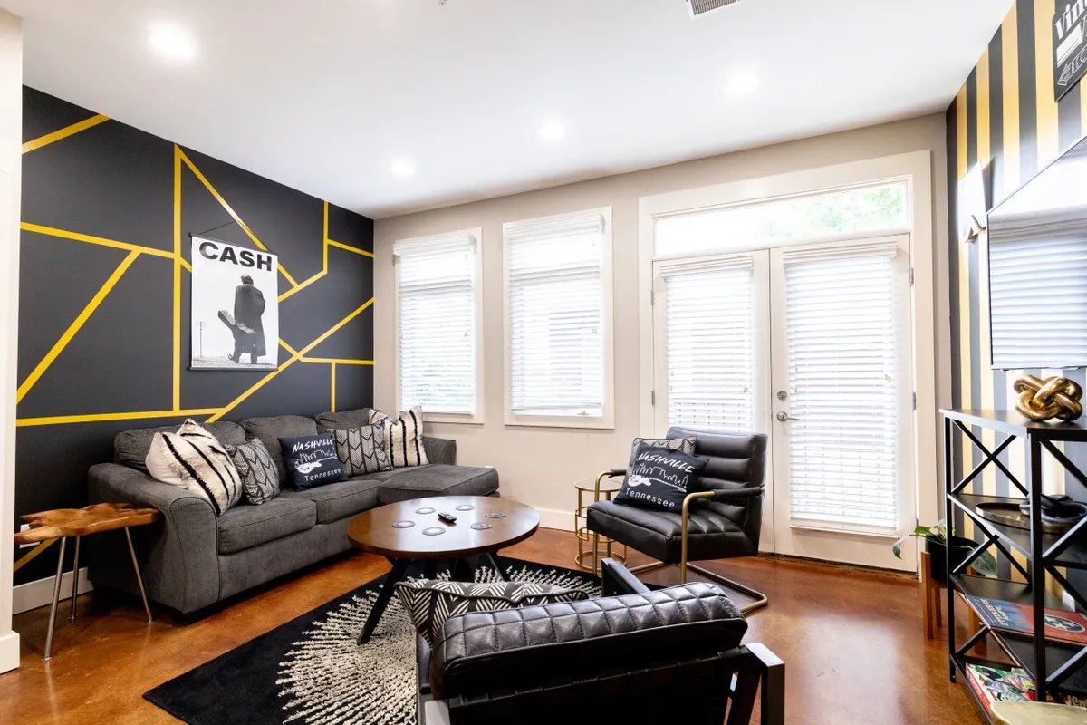 A bright, open living area featuring a modern geometric accent wall, contemporary sofa with decorative pillows, a sleek coffee table, and a black leather chair. The space is designed for relaxed social gatherings, with ample natural light and stylish decor that invites group interaction.