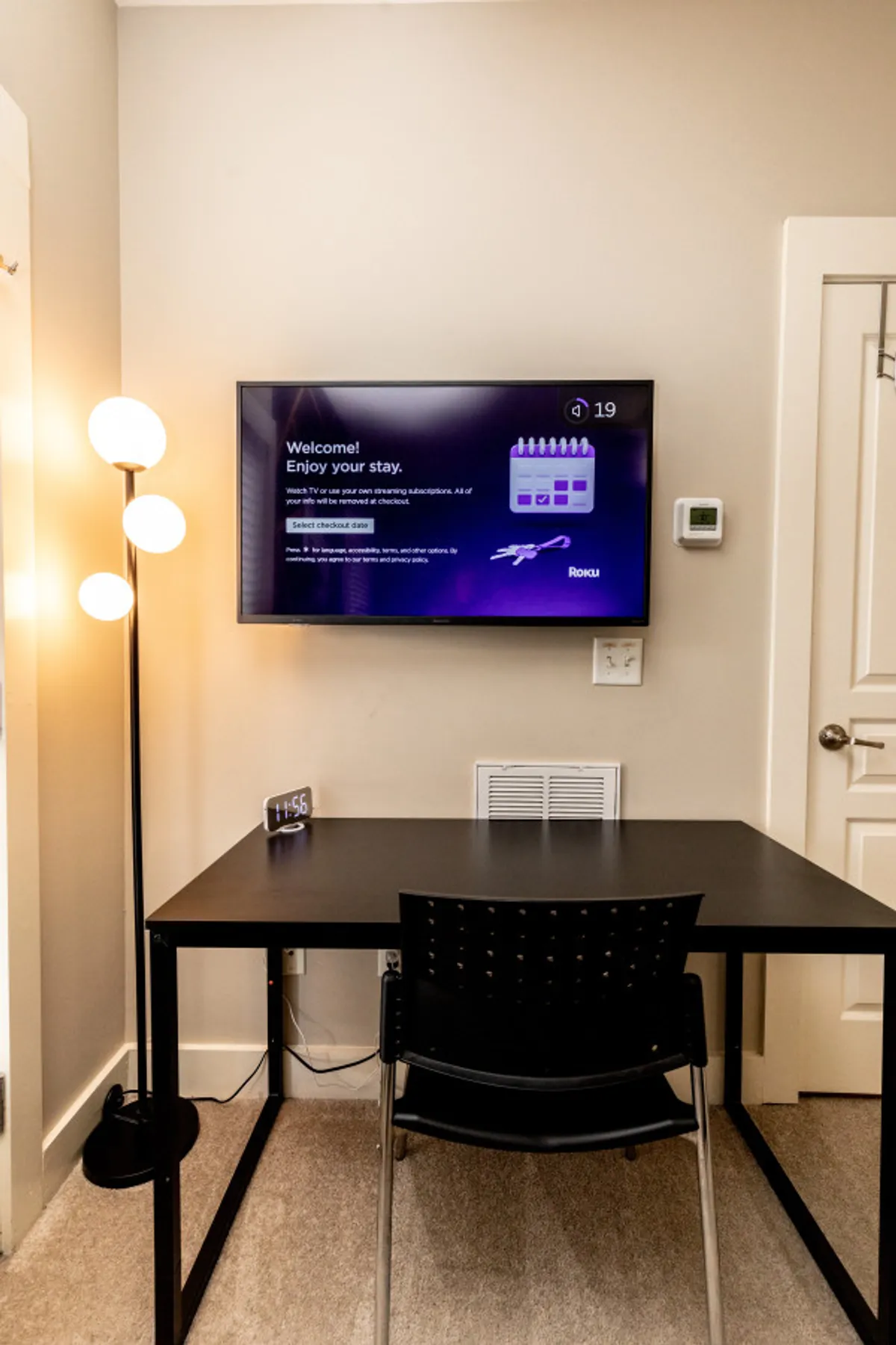 A modern living area featuring a television with a welcome message, a sleek floor lamp, a simple desk and chair, and a digital clock. The arrangement creates a cozy corner suitable for both work and relaxation, with thoughtful lighting and contemporary decor.